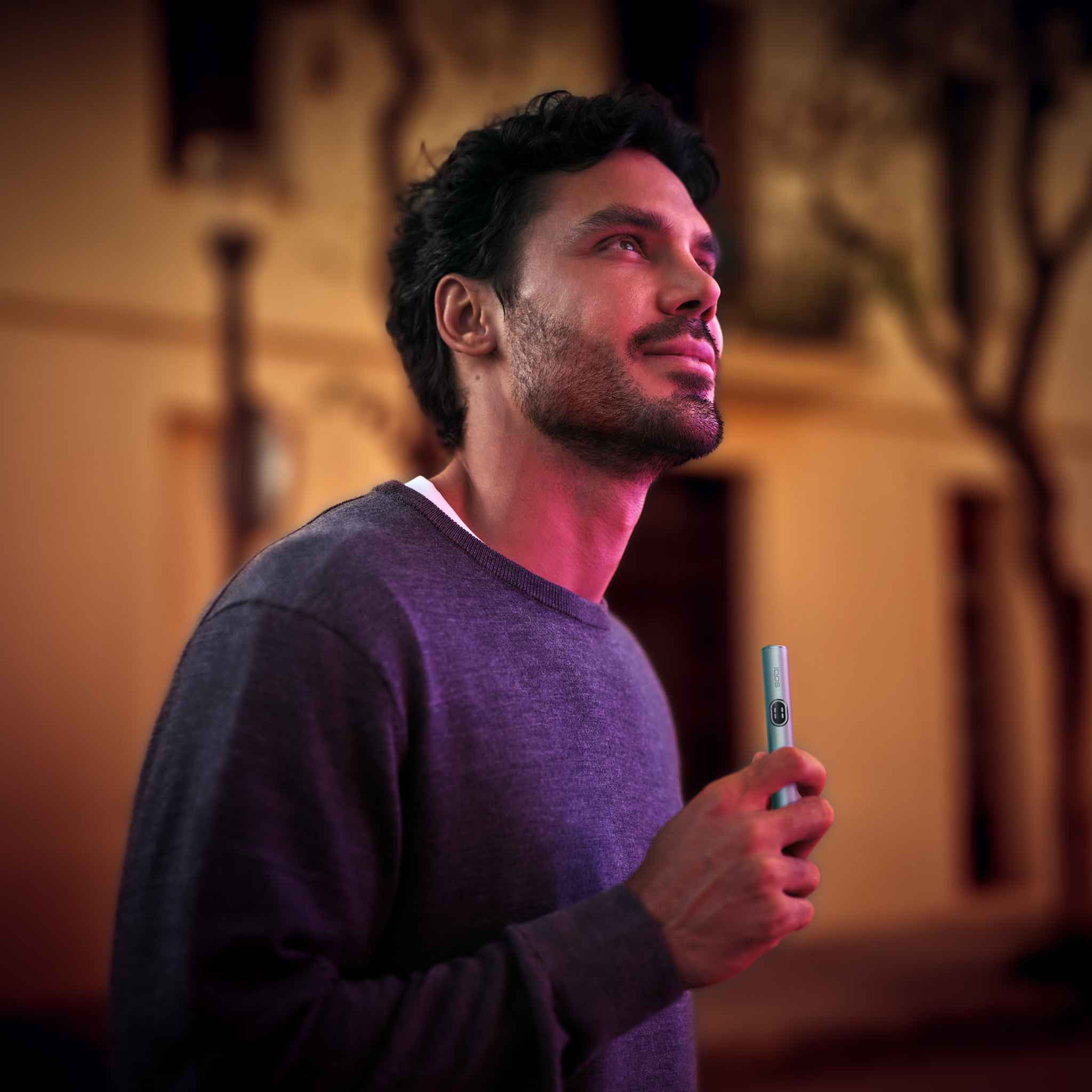 A person standing outside at night, holding an IQOS heated tobacco device. The background shows a blurred building and trees.
